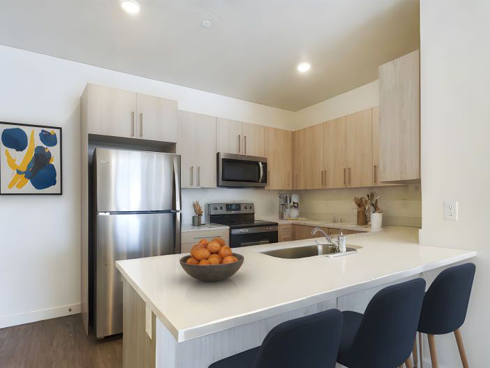 Modern kitchen in Apartments in Ogden featuring light wood cabinets, stainless steel appliances, a white island with sink, four dark stools, and a bowl of oranges on the counter. Abstract art adds a stylish touch to the space.
