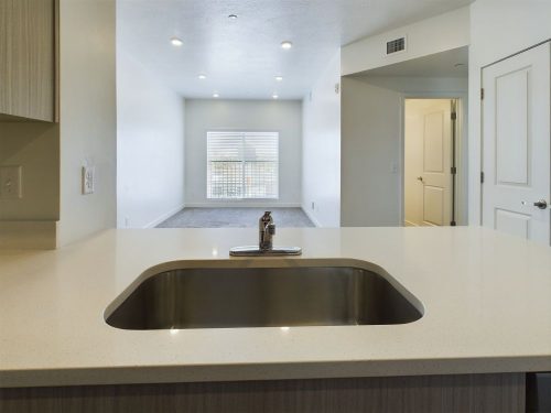 A modern kitchen sink with a stainless steel faucet on a quartz countertop overlooks an empty, well-lit living room with a large window.