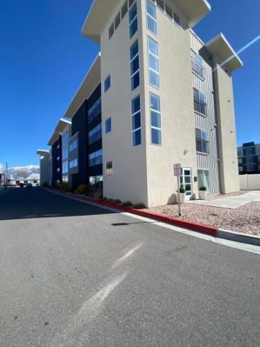 A modern multi-story apartment building with large windows, situated along a paved street under a clear blue sky.
