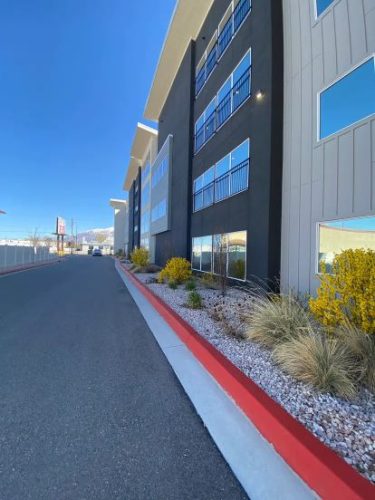 A modern, multi-story building with large windows, landscaped with rocks and shrubs alongside a paved road with a red-painted curb under a clear blue sky.