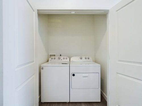 A white washing machine and dryer are placed side by side in a small laundry closet with double doors.