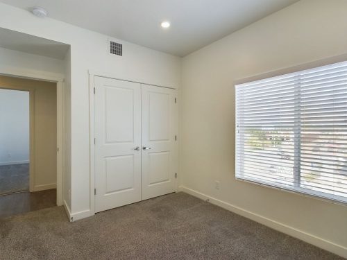 Empty bedroom with beige carpet, white walls, a double-door closet, a window with blinds, and an open doorway leading to another room.