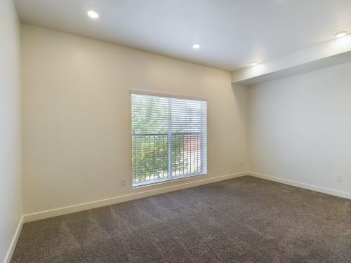 Empty room with beige walls, gray carpet, recessed ceiling lights, and a large window with blinds letting in natural light.