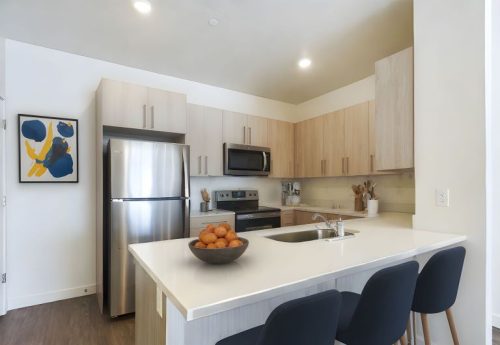 Modern kitchen with light wood cabinets, stainless steel appliances, a bowl of oranges on the counter, and three black barstools at the island.