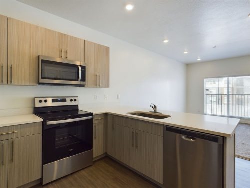 Modern kitchen with light wood cabinets, stainless steel appliances including a stove, microwave, and dishwasher, white countertops, and an open layout with bright natural light.