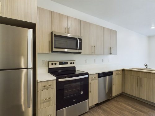 Modern kitchen with light wood cabinets, stainless steel refrigerator, stove, microwave, dishwasher, and a sink in the corner.